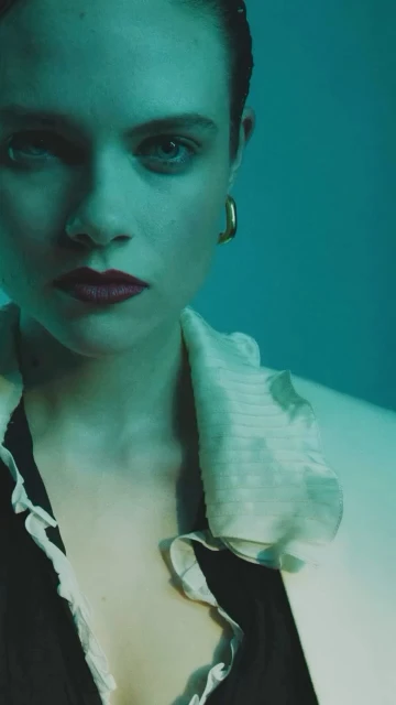 Portrait of a young woman with red lipstick and gold earrings, wearing a black top with a white ruffled collar, against a teal background.