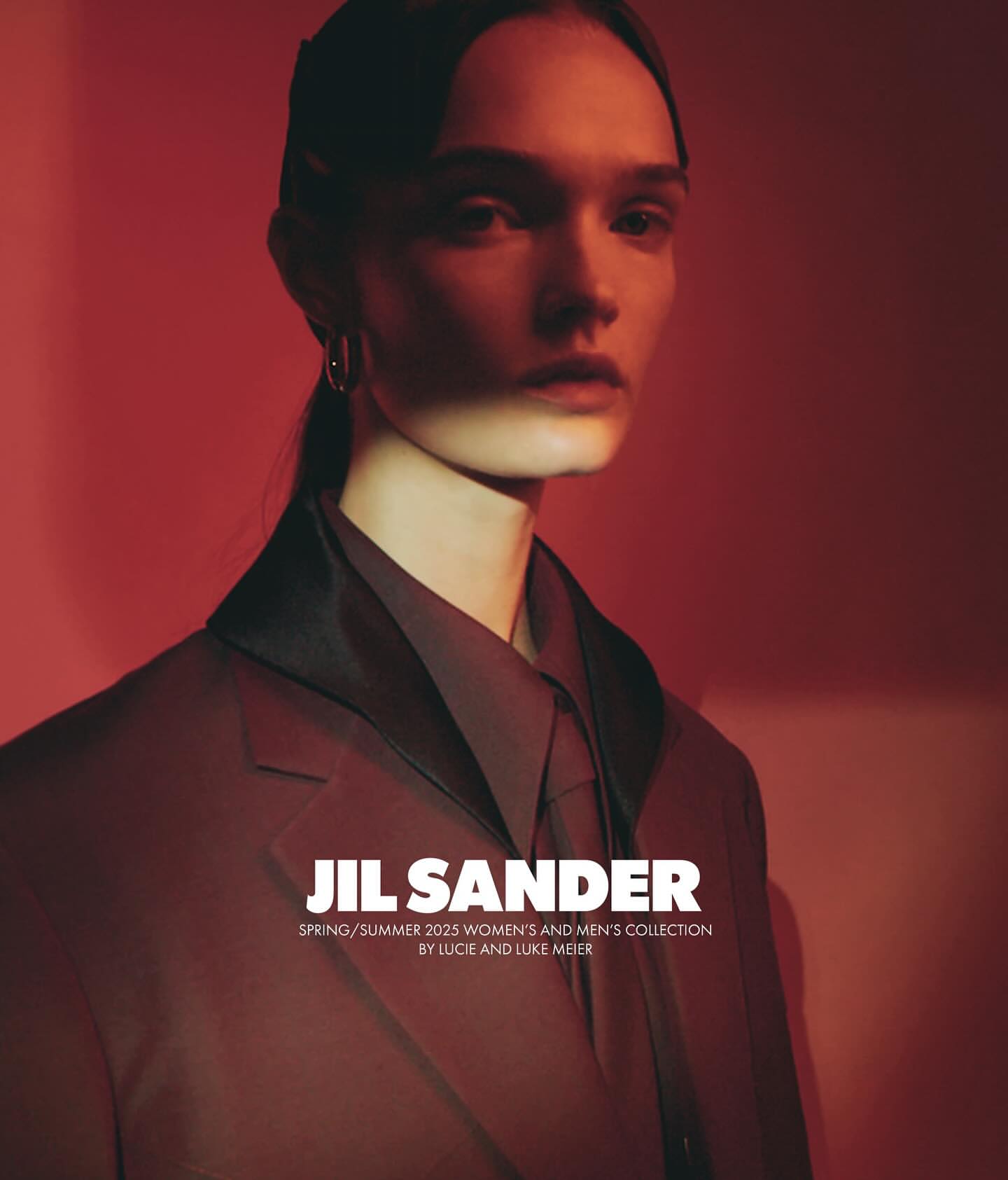 Fashion editorial shot of a fair-skinned adult female model in a brown suit, posed in a photo studio with a vibrant red backdrop and light.