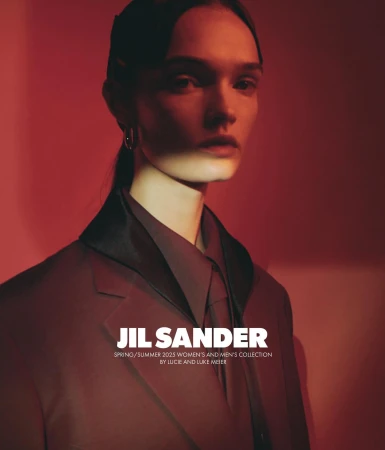 Fashion editorial shot of a fair-skinned adult female model in a brown suit, posed in a photo studio with a vibrant red backdrop and light.