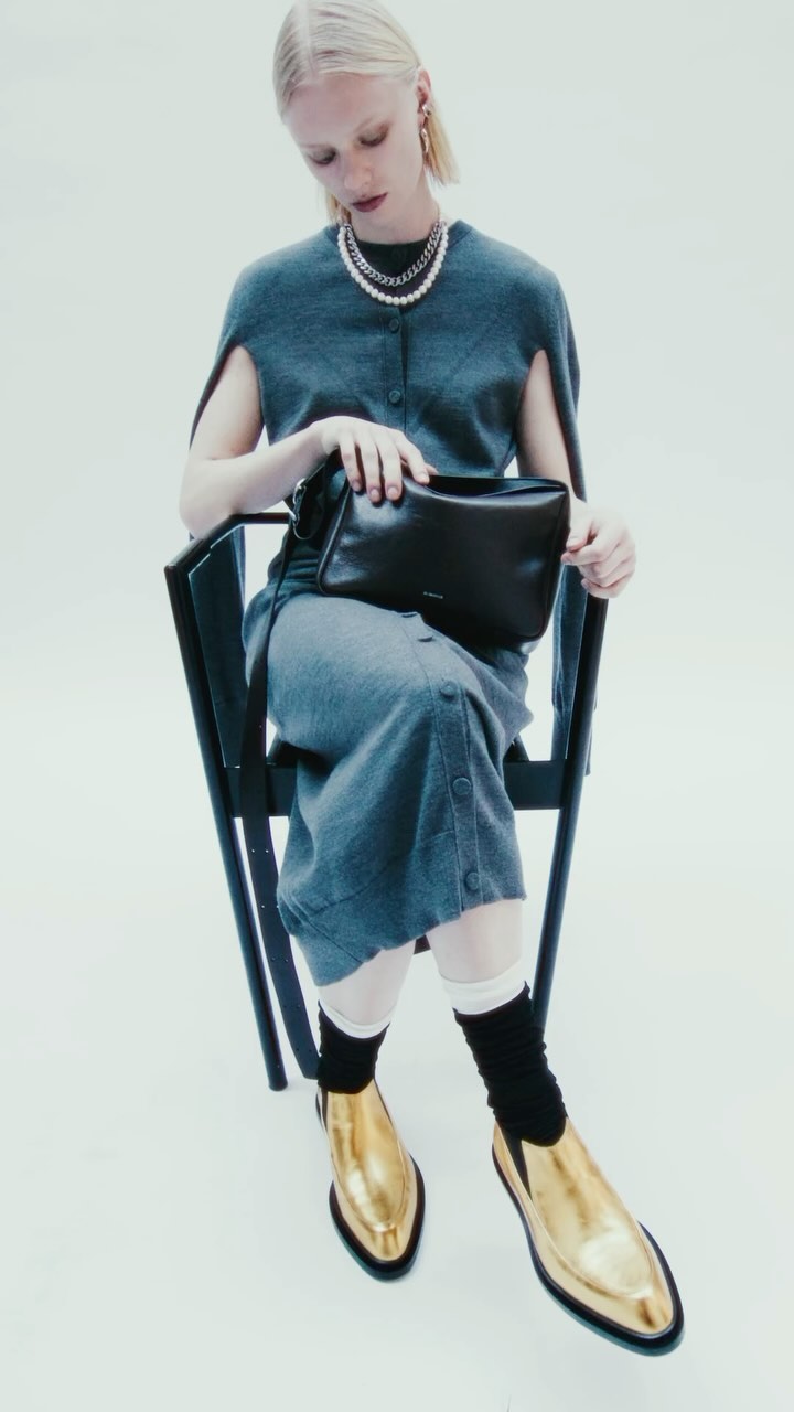 Fashion shot of a woman in a grey outfit sitting on a chair with a black purse and gold shoes against a white background.