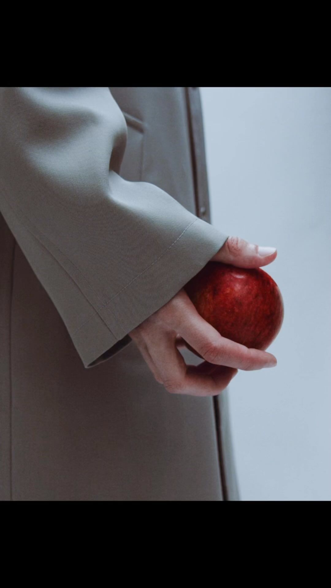 Close-up of an adult's hand holding a red apple while wearing a beige trench coat in a brightly lit studio environment.