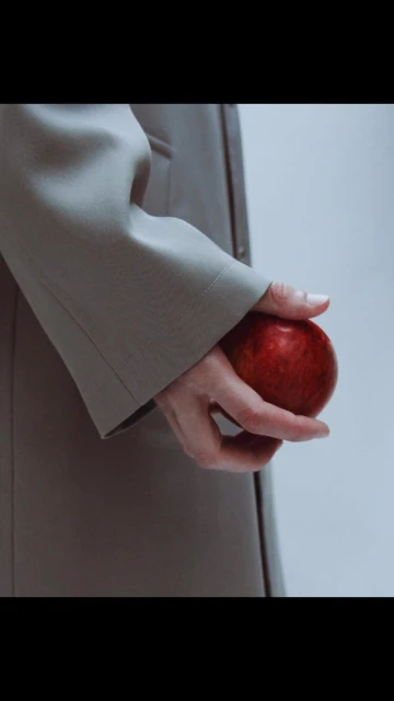 Close-up of an adult's hand holding a red apple while wearing a beige trench coat in a brightly lit studio environment.