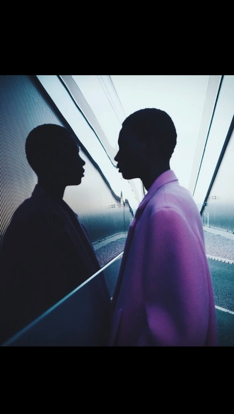Woman in a pink blazer stands in front of an escalator with a mirrored wall, reflecting her silhouette in an indoor setting.