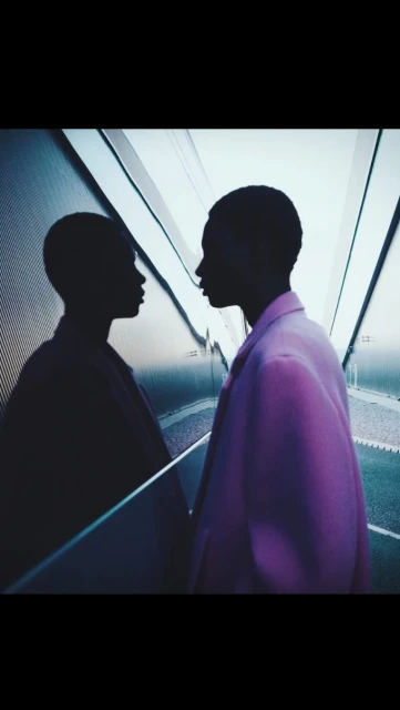 Woman in a pink blazer stands in front of an escalator with a mirrored wall, reflecting her silhouette in an indoor setting.