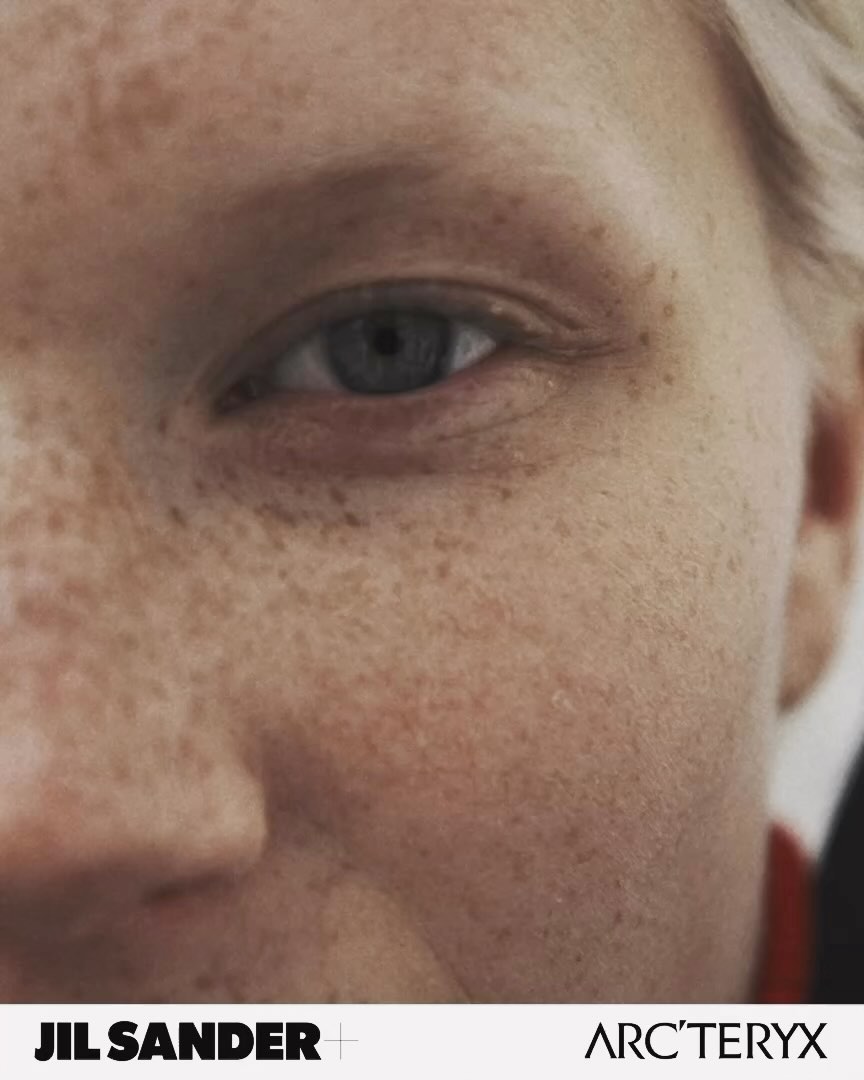 Close-up of an adult female's face, showcasing freckled skin and a striking blue eye, with brand logos JIL SANDER and ARC'TERYX visible.