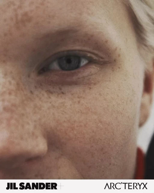 Close-up of an adult female's face, showcasing freckled skin and a striking blue eye, with brand logos JIL SANDER and ARC'TERYX visible.