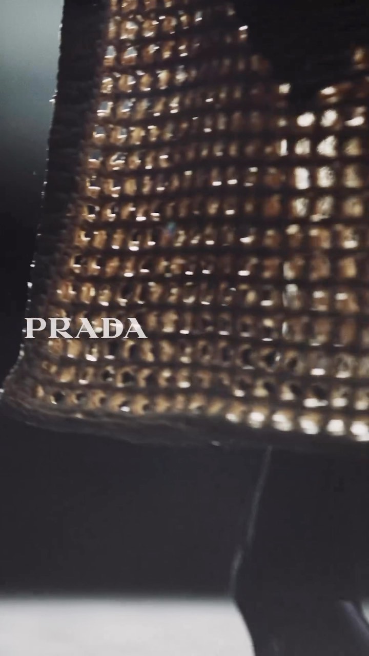 Close-up fashion shot of a Prada dress with metallic material and patterned design, showcasing the brand's logo and texture against a dark background.