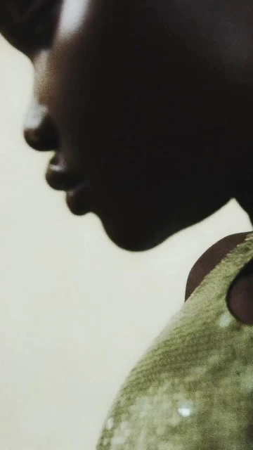 Close-up profile of a Black female adult with a sequined garment. Dark complexion, neutral expression, facing left against a neutral background.