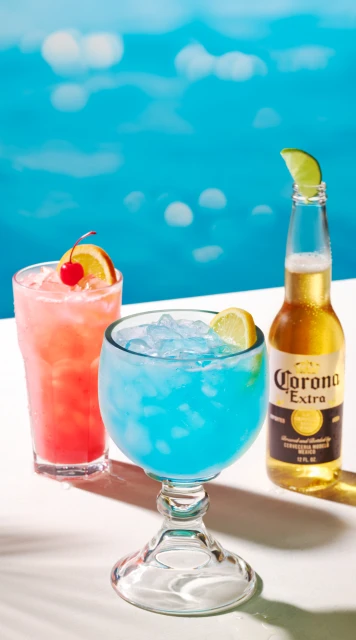 Three beverages including a bright blue cocktail, a pink cocktail, and a bottle of beer are staged against a blurred blue background.