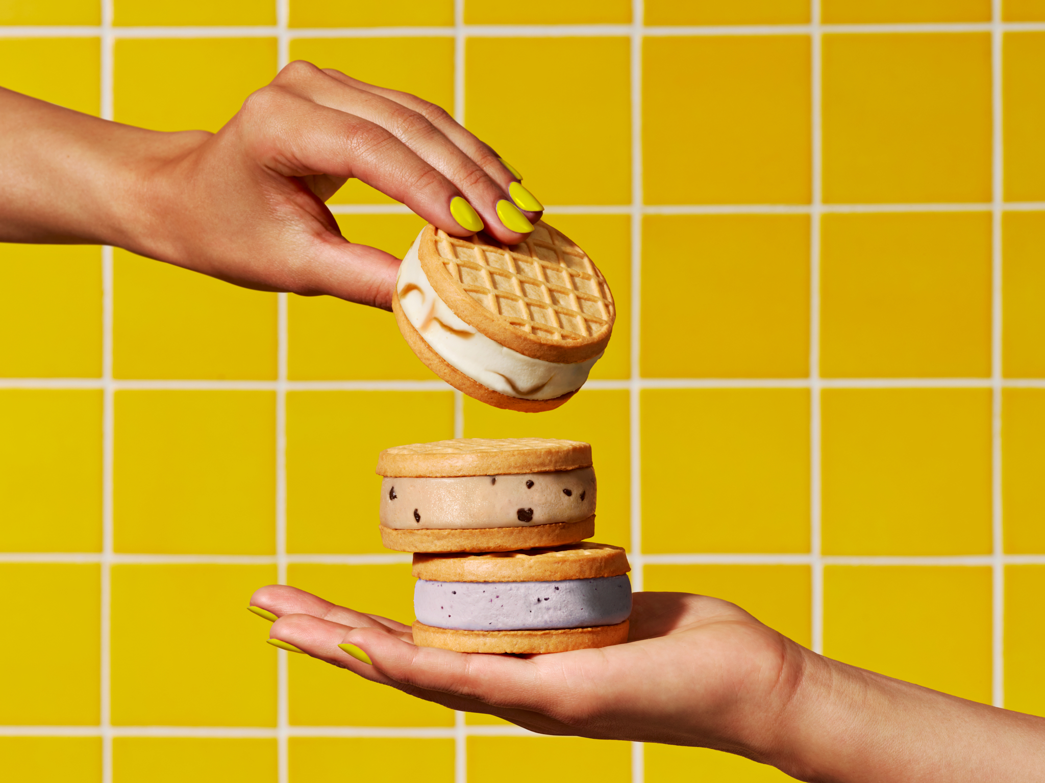 Two hands present a tower of ice cream cookie sandwiches with waffle cone cookies against a bright yellow tiled background.