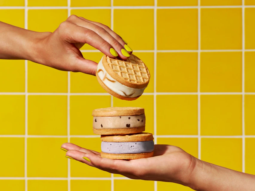 Two hands present a tower of ice cream cookie sandwiches with waffle cone cookies against a bright yellow tiled background.
