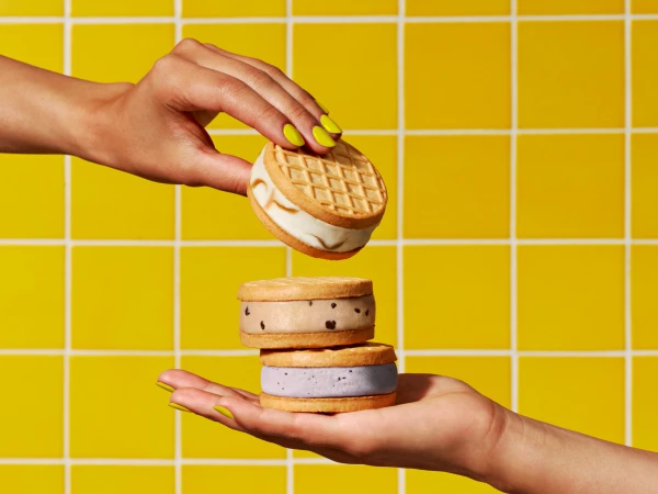 Two hands present a tower of ice cream cookie sandwiches with waffle cone cookies against a bright yellow tiled background.