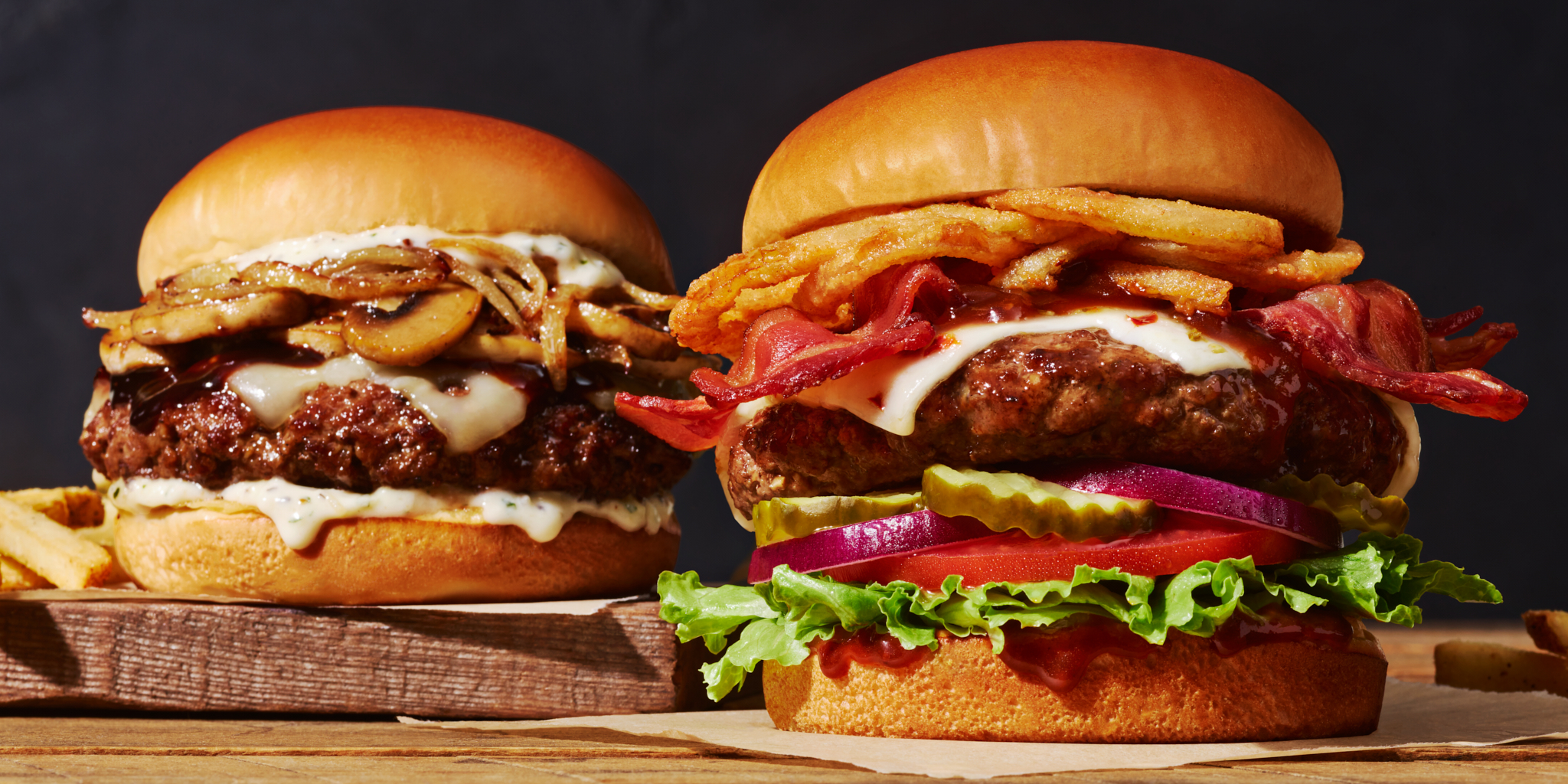 Two gourmet hamburgers with toppings like bacon, onion rings, mushrooms, and various vegetables served on a wooden board.
