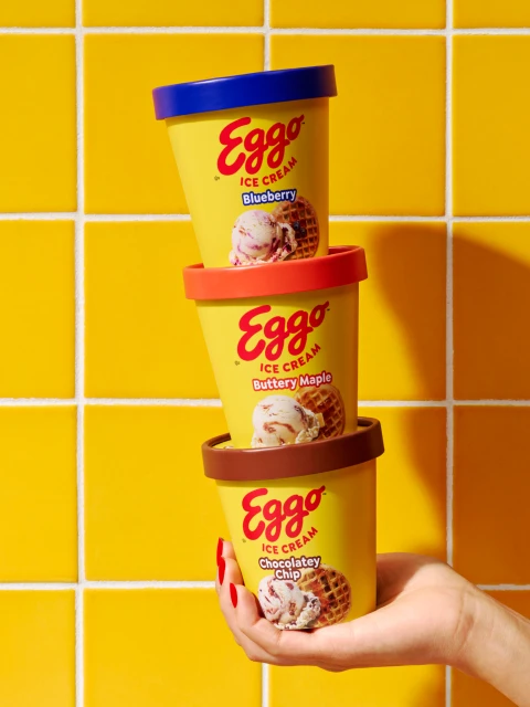Stack of three Eggo ice cream containers in blueberry, buttery maple, and chocolatey chip flavors held by a person with red nail polish.