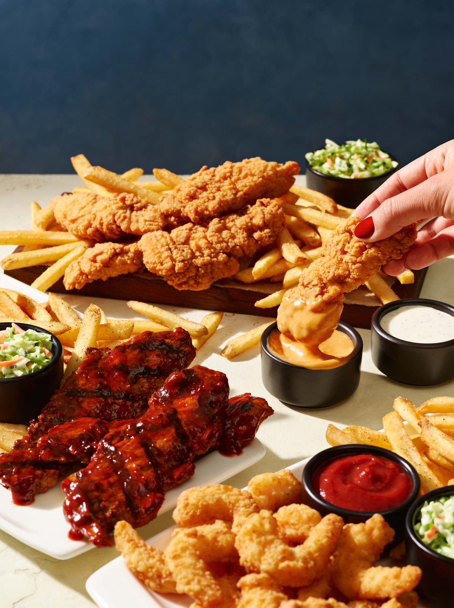 A person dunks a crispy fried chicken strip into a creamy sauce, amidst a hearty spread of ribs, shrimp, fries, and coleslaw.