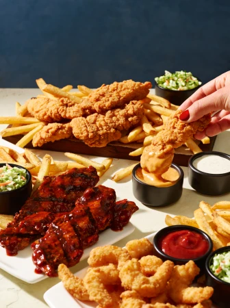 A person dunks a crispy fried chicken strip into a creamy sauce, amidst a hearty spread of ribs, shrimp, fries, and coleslaw.