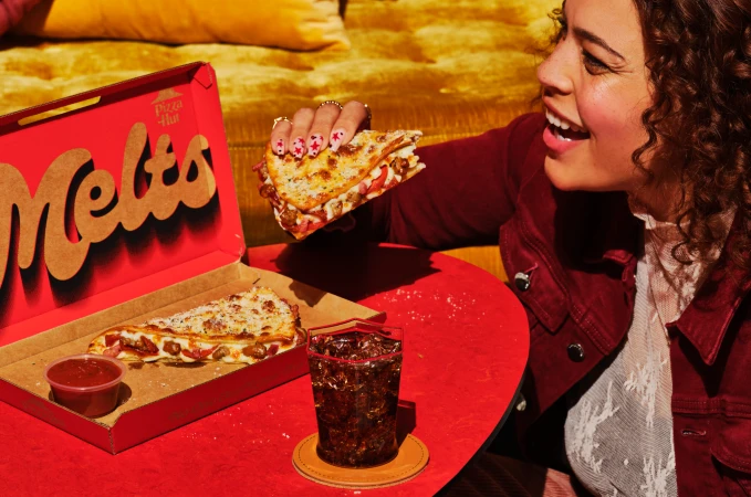 Smiling woman holds Pizza Hut Melt sandwich with a box, soda, and dipping sauce on a red table against a yellow-gold background.