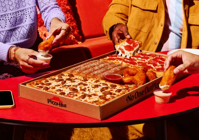 Four people gather around a red table to enjoy a variety of pizzas, chicken wings, and dipping sauces from a Pizza Hut party box.