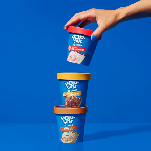 Three vibrant Pop-Tarts ice cream pints stacked against a bright blue backdrop, showcasing strawberry, s'mores, and brown sugar cinnamon flavors.