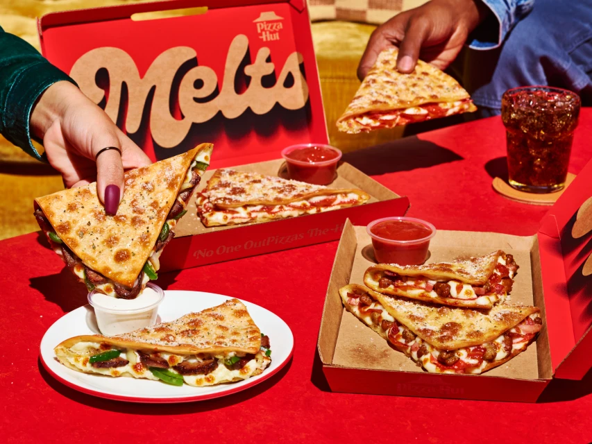 Overhead shot of Pizza Hut melts, served in open cardboard boxes and on a plate, presented on a red table, with two people visible.