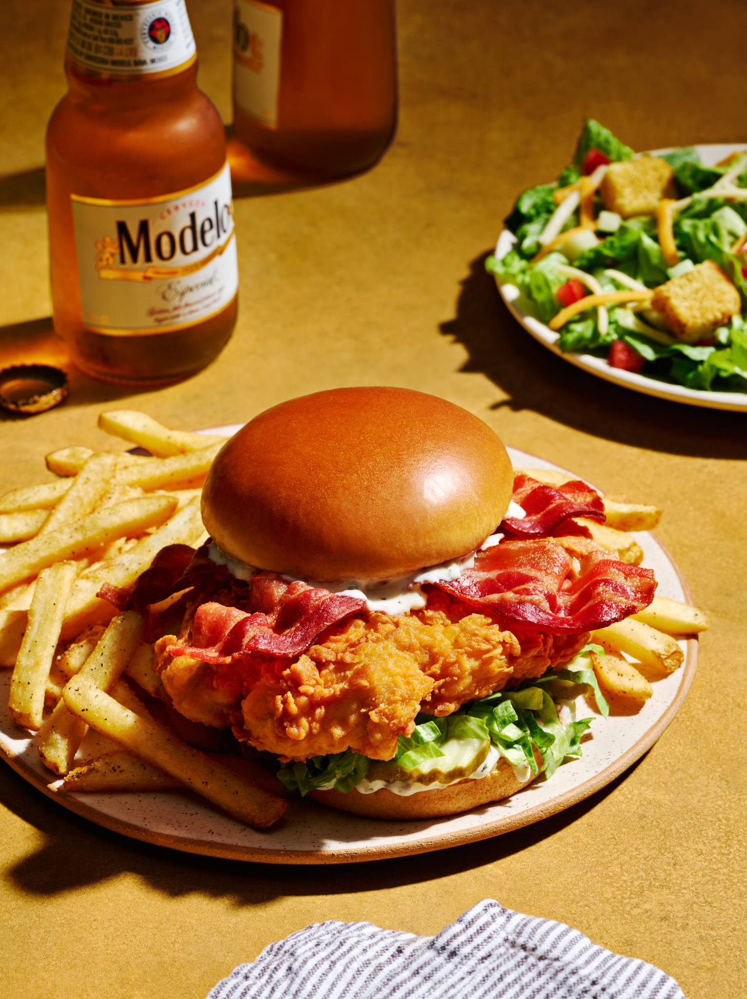 Crispy chicken sandwich with bacon, lettuce, and fries on a plate, accompanied by a bottle of Modelo beer and a side salad.