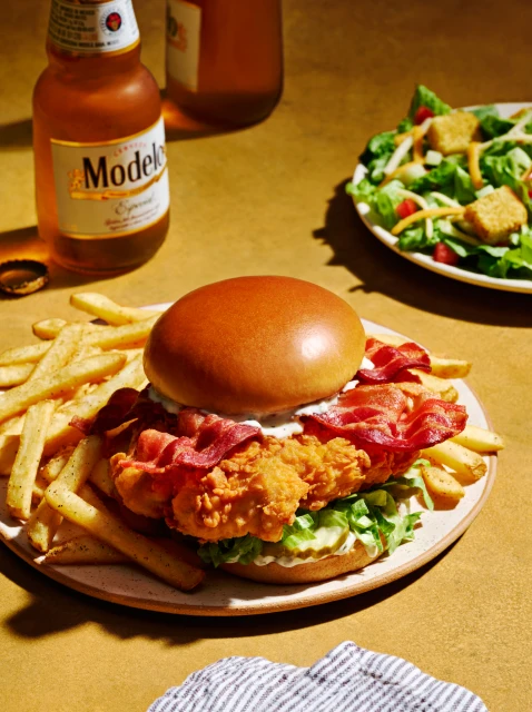 Crispy chicken sandwich with bacon, lettuce, and fries on a plate, accompanied by a bottle of Modelo beer and a side salad.