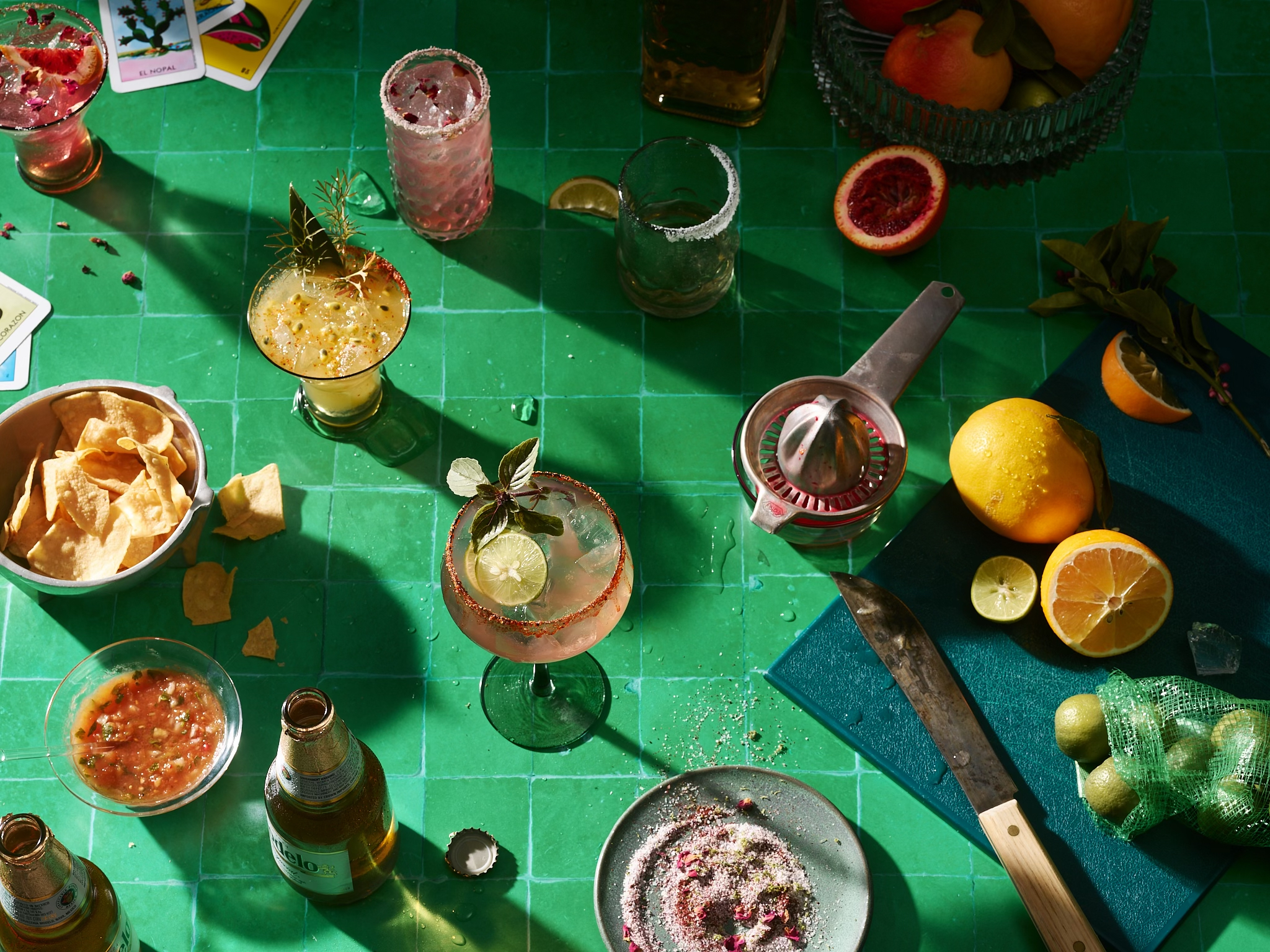 Overhead shot of a brightly lit green tiled surface with a variety of cocktails, snacks, citrus fruit, and Mexican beer bottles.