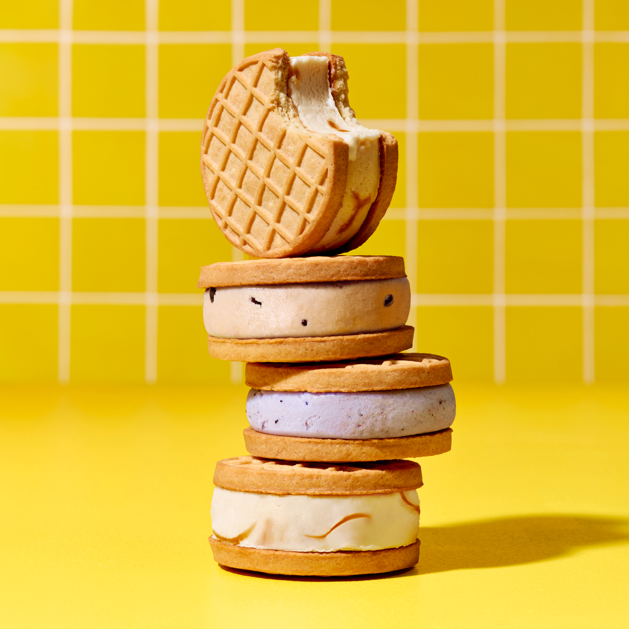 A stack of ice cream sandwiches with waffle cookies sits in front of a yellow grid background, creating a sweet and visually appealing dessert image.