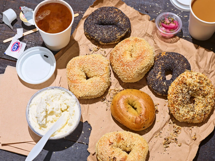 Overhead shot of assorted bagels with cream cheese and coffee on craft paper, creating a vibrant, appealing food still life.
