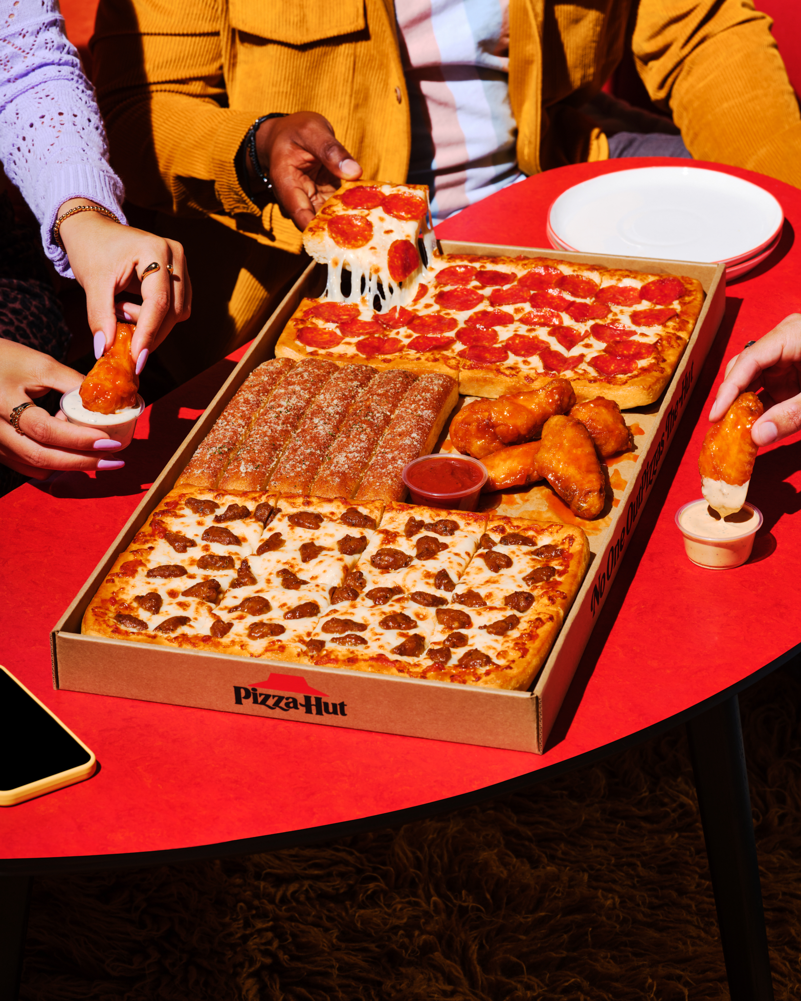 People share a Pizza Hut Big Dinner Box on a red table. The box includes pepperoni pizza, sausage pizza, breadsticks, and chicken wings.