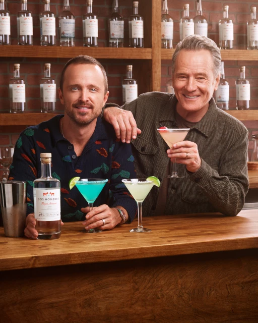 Bryan Cranston and Aaron Paul pose behind a bar with cocktails and a bottle of Dos Hombres Mezcal, showcasing the product.