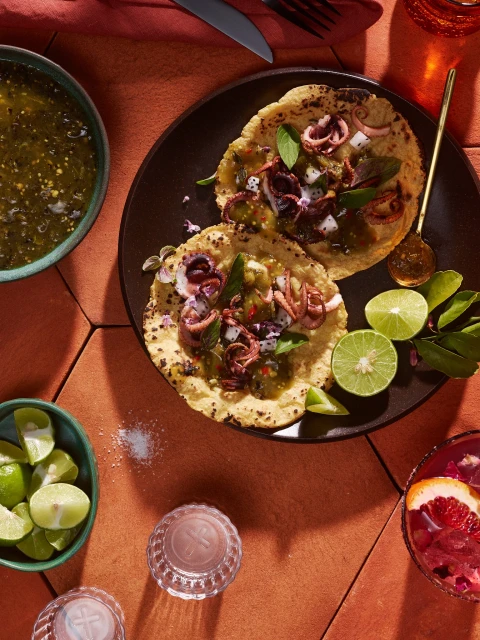 Overhead shot of two octopus tacos on a black plate surrounded by lime wedges, green salsa, and grapefruit cocktail on a terracotta surface.