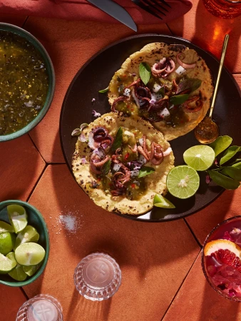 Overhead shot of two octopus tacos on a black plate surrounded by lime wedges, green salsa, and grapefruit cocktail on a terracotta surface.