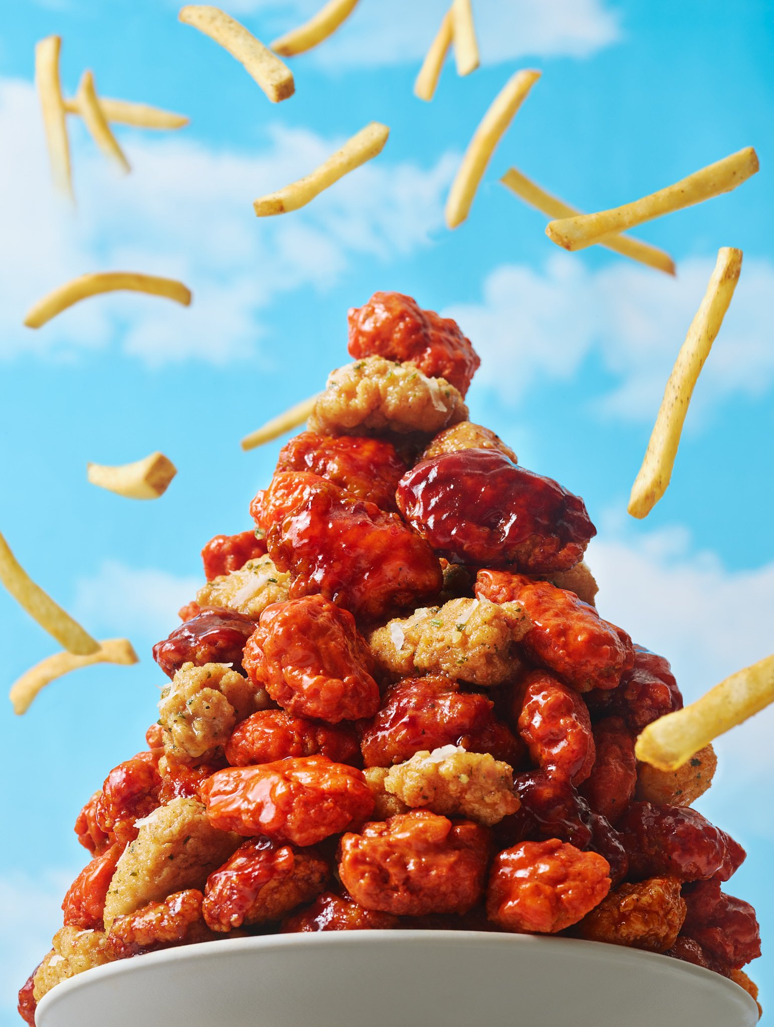 Overhead shot of a mountain of saucy chicken wings and fries in a white bowl against a blue sky with scattered clouds background.