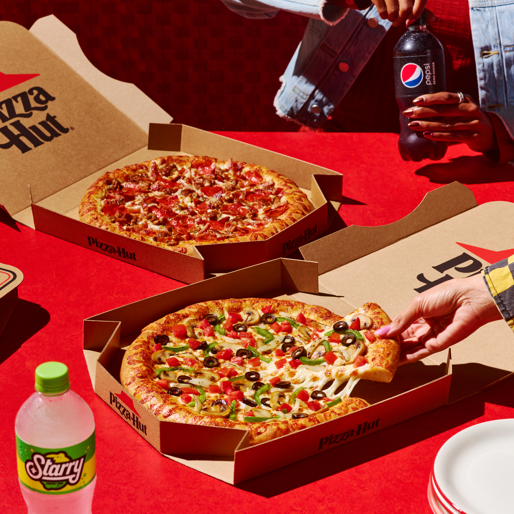 Two Pizza Hut pizzas in boxes, one with a slice being lifted, soda bottles, and plates are arranged on a red surface.