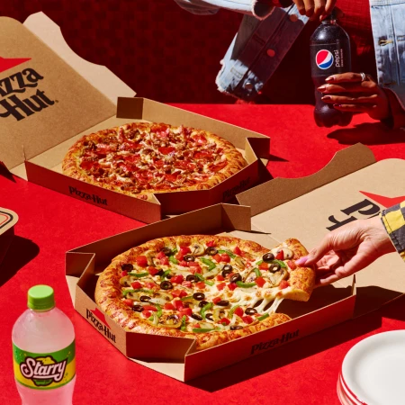 Two Pizza Hut pizzas in boxes, one with a slice being lifted, soda bottles, and plates are arranged on a red surface.