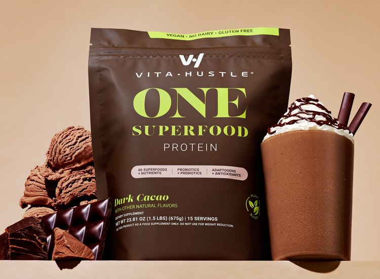 Vita Hustle One Superfood protein powder in Dark Cacao flavor, accompanied by a chocolate protein shake and chocolate pieces on a tan surface.