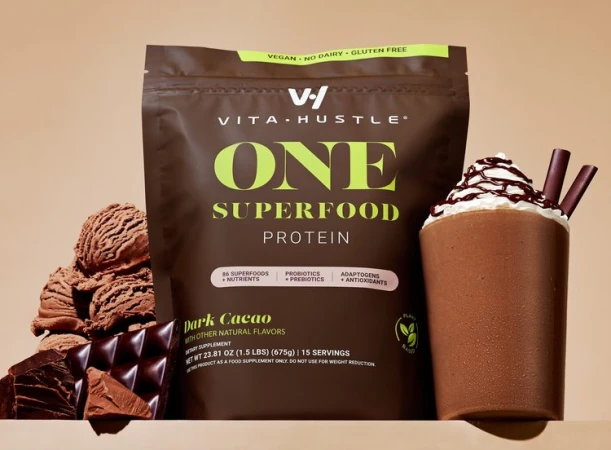 Vita Hustle One Superfood protein powder in Dark Cacao flavor, accompanied by a chocolate protein shake and chocolate pieces on a tan surface.