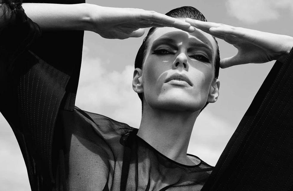 Monochrome portrait of a woman with hands above her forehead, dark eye makeup, wearing a sheer top, against a sky background.