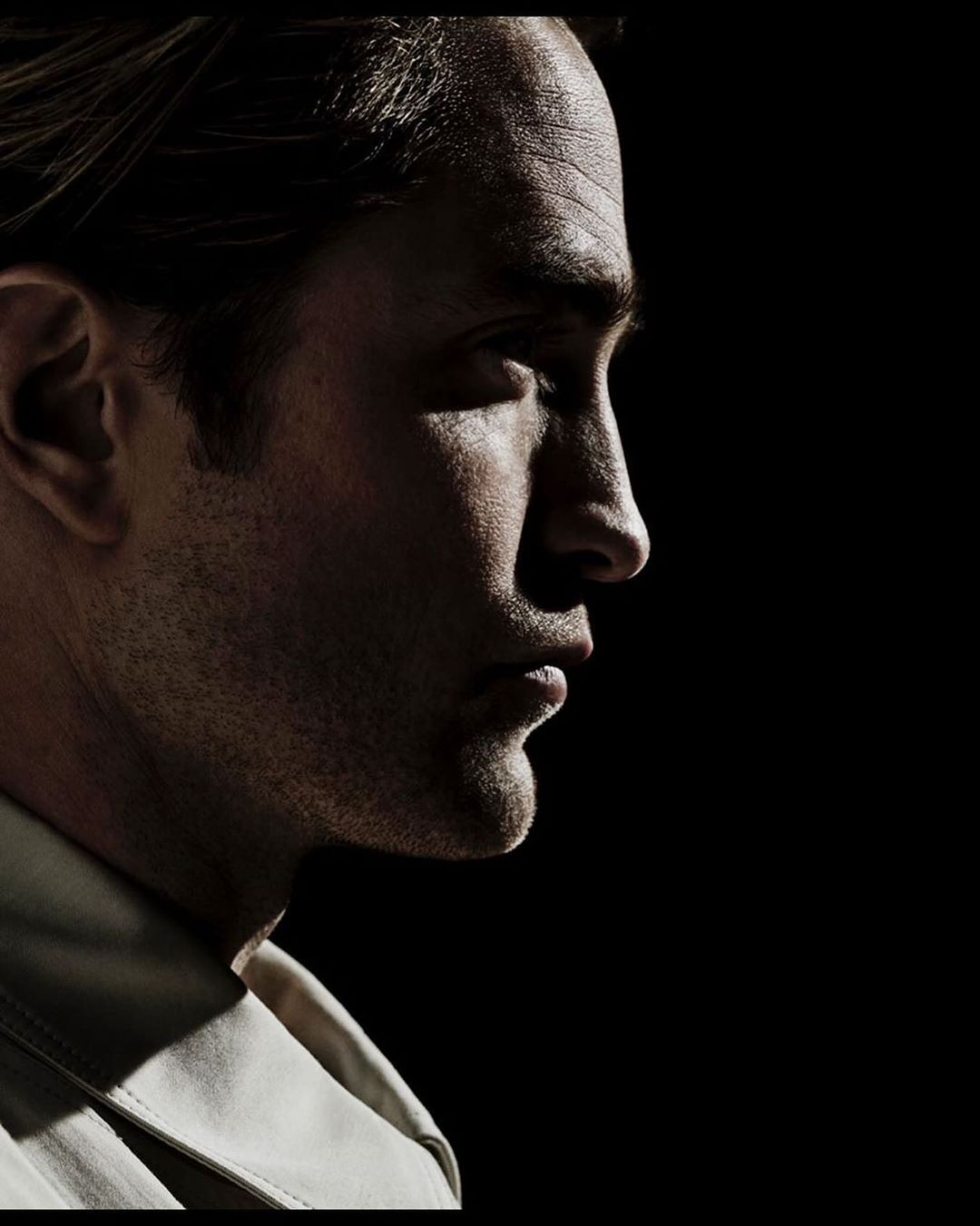 Dramatic portrait of a man in profile with strong lighting, showcasing his facial features against a stark black backdrop, wearing a light-colored shirt.