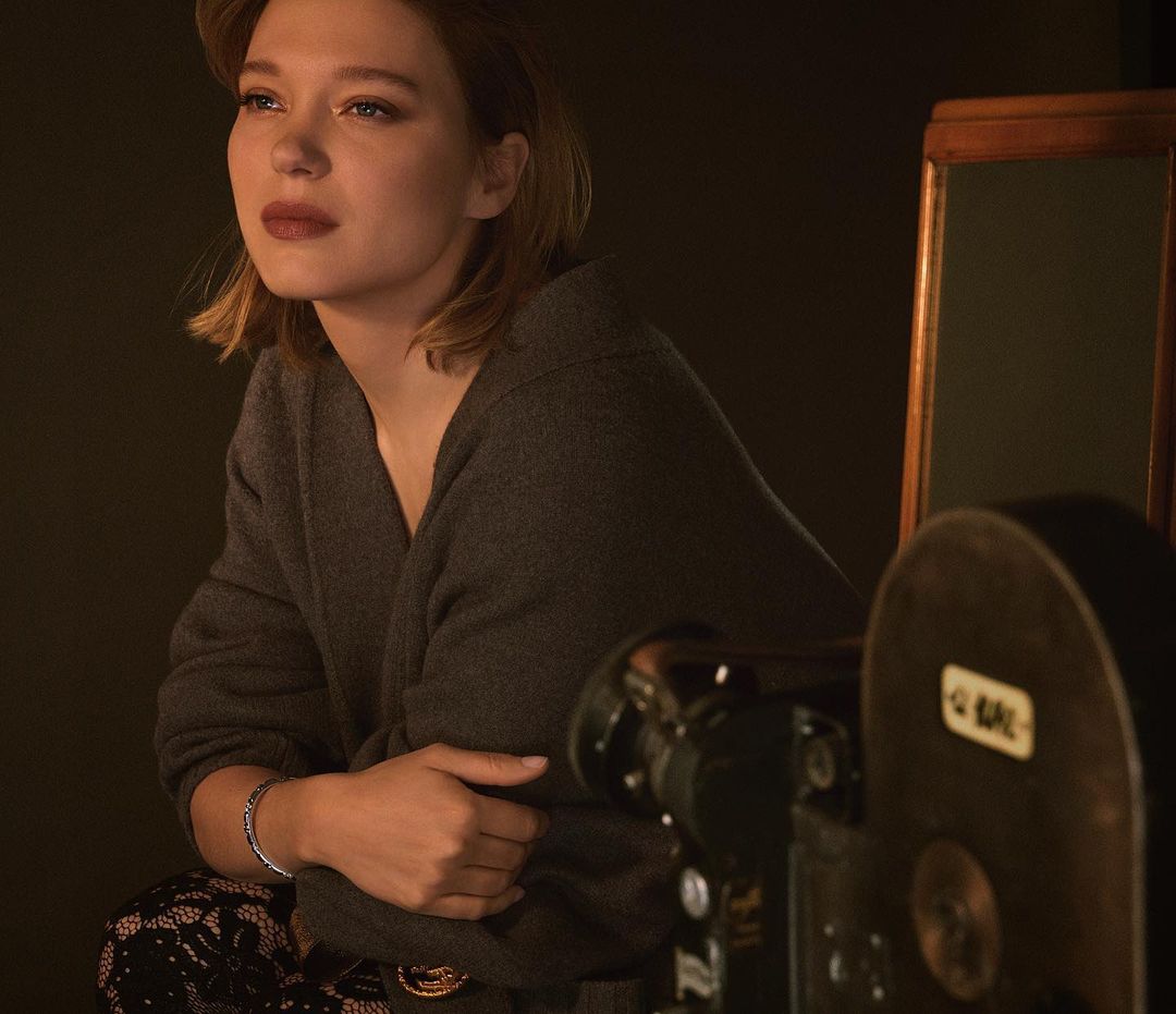 Portrait of a blonde woman wearing a gray sweater posing in front of a vintage camera and dark backdrop, with a contemplative expression.