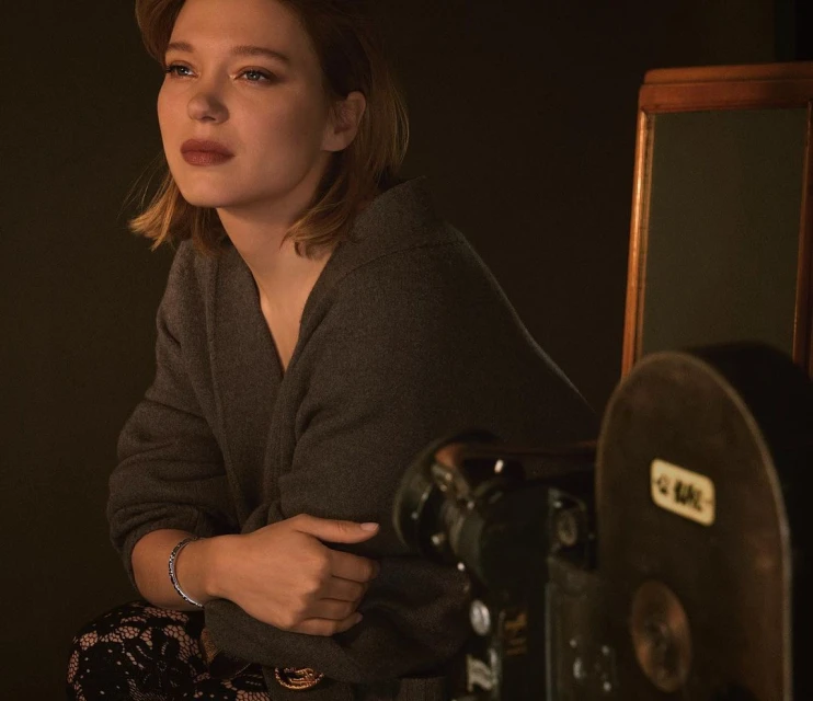 Portrait of a blonde woman wearing a gray sweater posing in front of a vintage camera and dark backdrop, with a contemplative expression.
