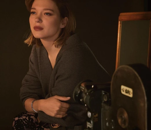 Portrait of a blonde woman wearing a gray sweater posing in front of a vintage camera and dark backdrop, with a contemplative expression.