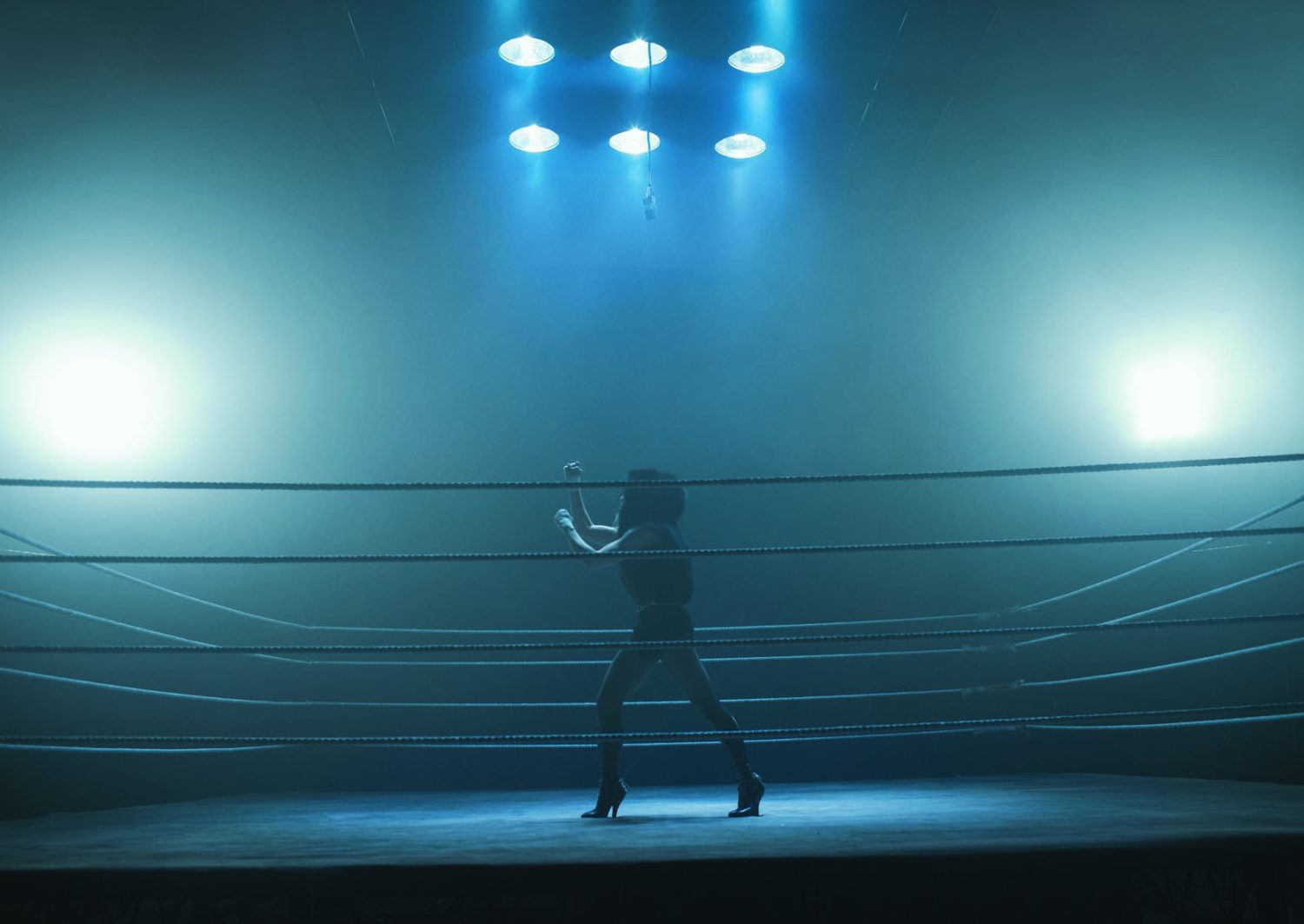 Female boxer in a fighting stance in a boxing ring, illuminated by bright stage lights, creating a dramatic, high-contrast scene.