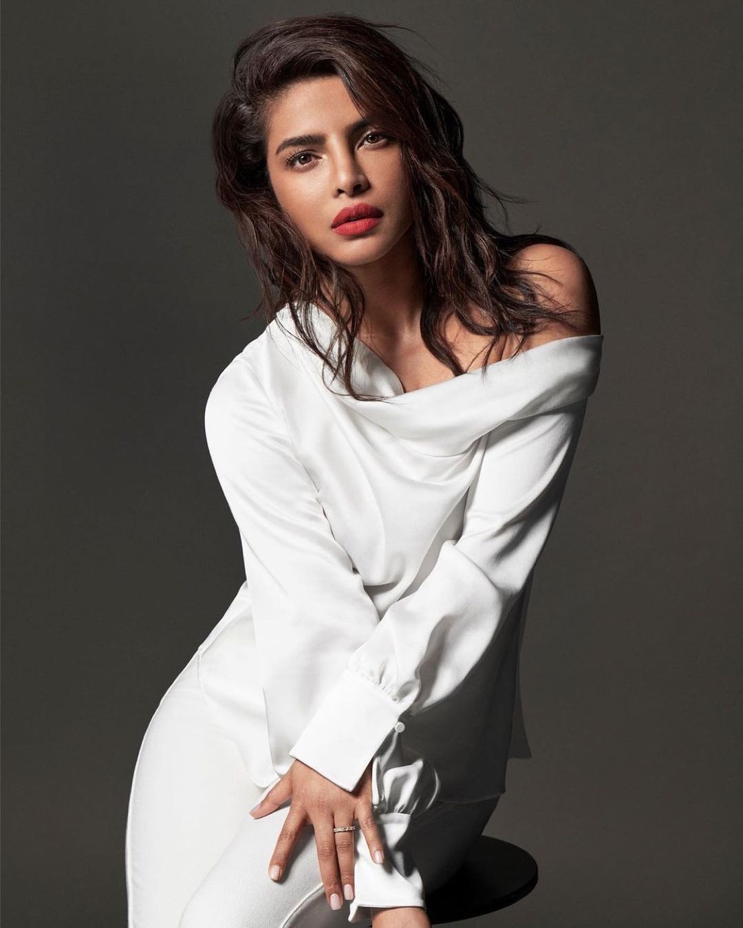 Portrait of a brunette adult woman in a white off-the-shoulder top and light-colored pants posing in front of a grey background.