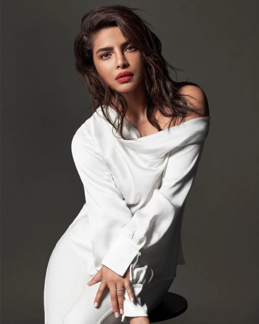 Portrait of a brunette adult woman in a white off-the-shoulder top and light-colored pants posing in front of a grey background.