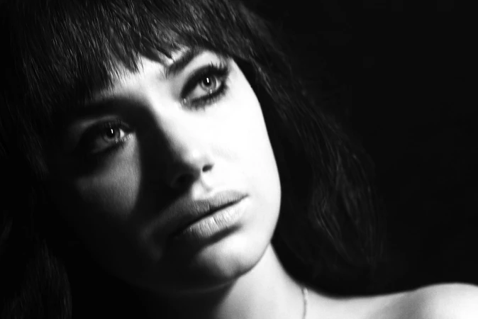 Close-up monochrome portrait of a woman with bangs and dark eye makeup looking upwards against a black background.