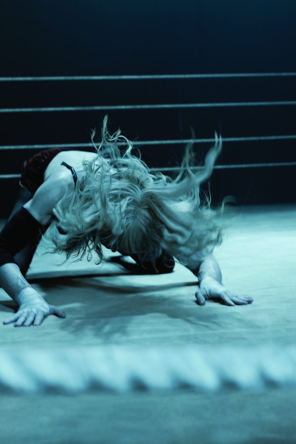 Female wrestler in a ring with blonde hair, positioned on the mat with her hands. She is wearing a dark top and arm bands.
