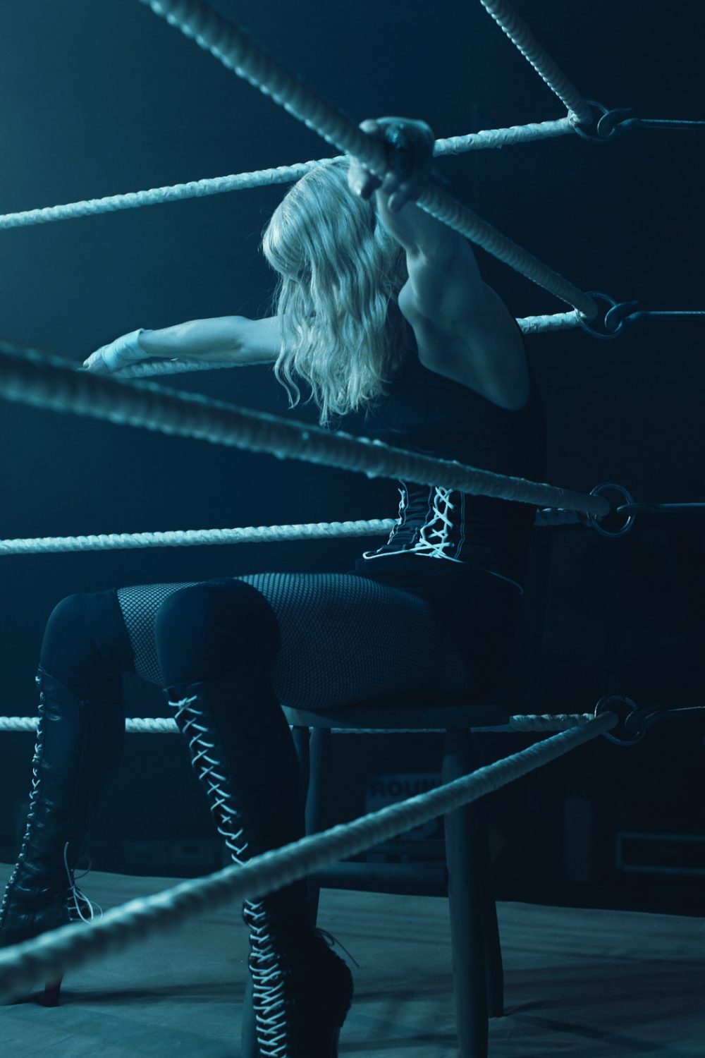 Woman sitting on a stool, leaning against the ropes in a dimly lit boxing ring, wearing a corset, fishnet stockings, and lace-up thigh-high boots.