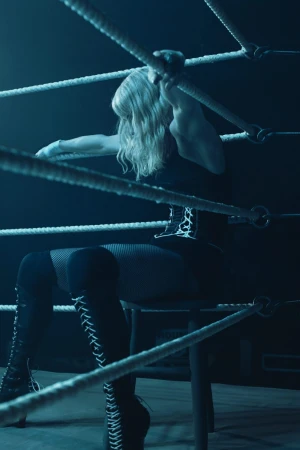 Woman sitting on a stool, leaning against the ropes in a dimly lit boxing ring, wearing a corset, fishnet stockings, and lace-up thigh-high boots.